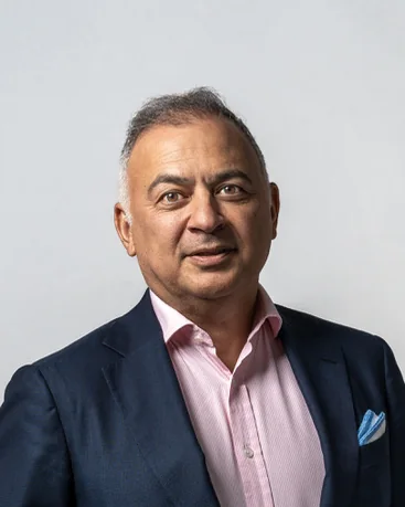 A man in a navy suit and pink shirt on a grey backdrop