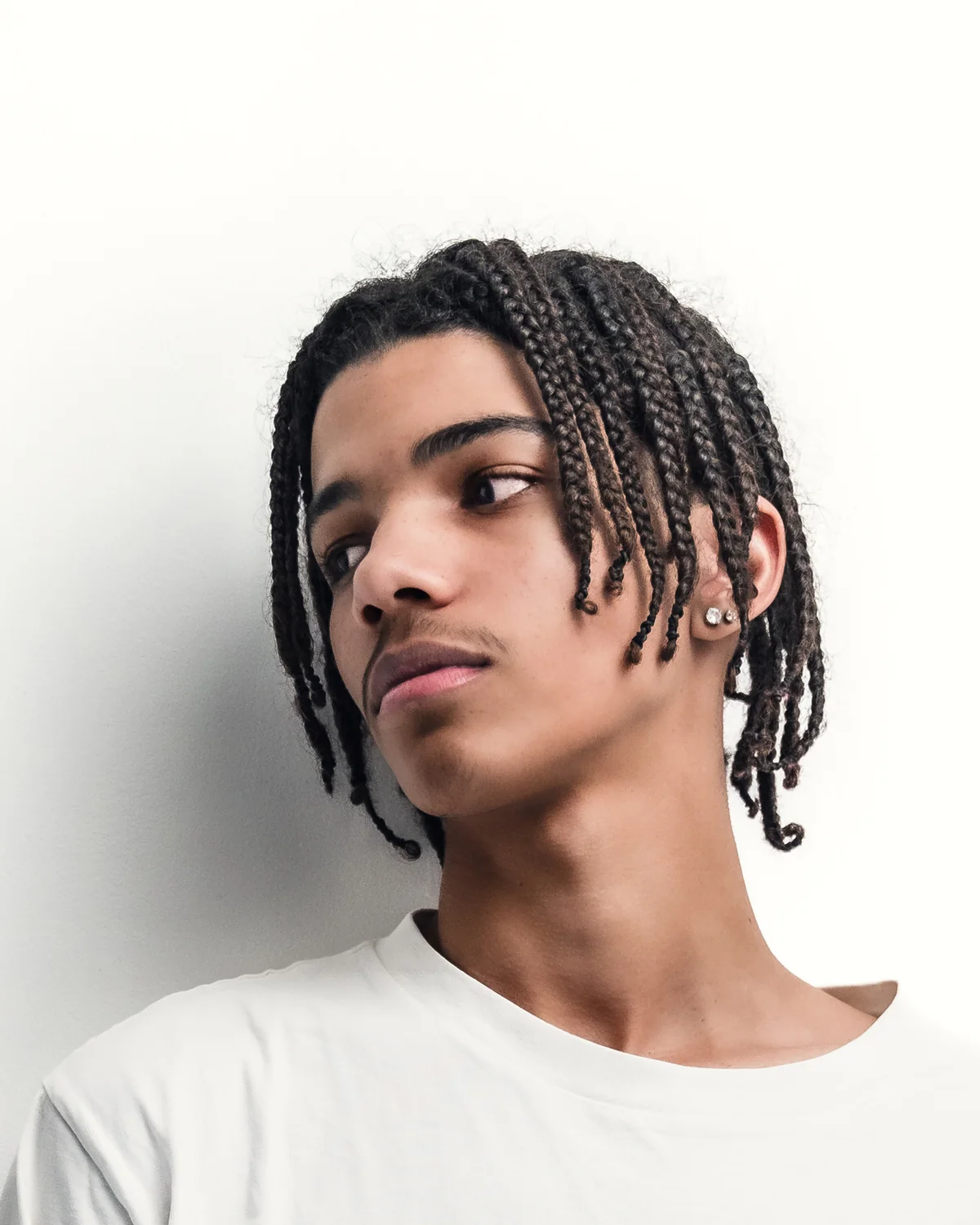 A young man with braids against a white wall, looking aside