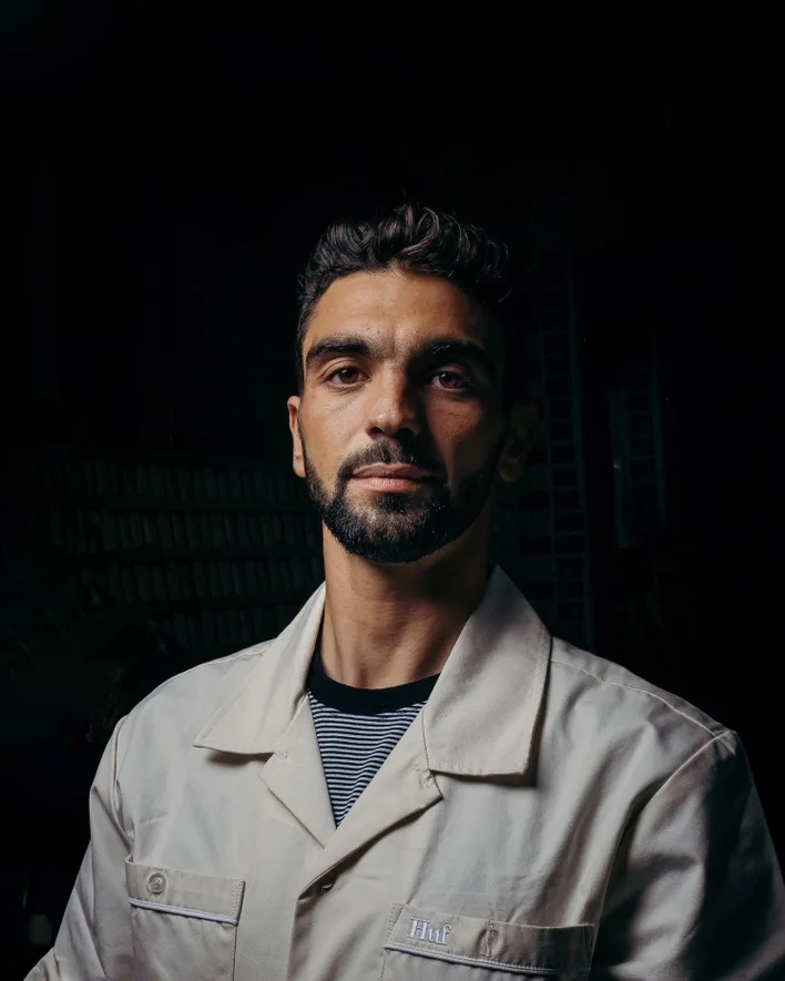 A man with a beard in a cream work shirt on a dark backdrop