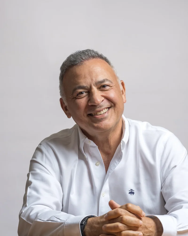 A smiling man in a white shirt, hands clasped, on a grey backdrop