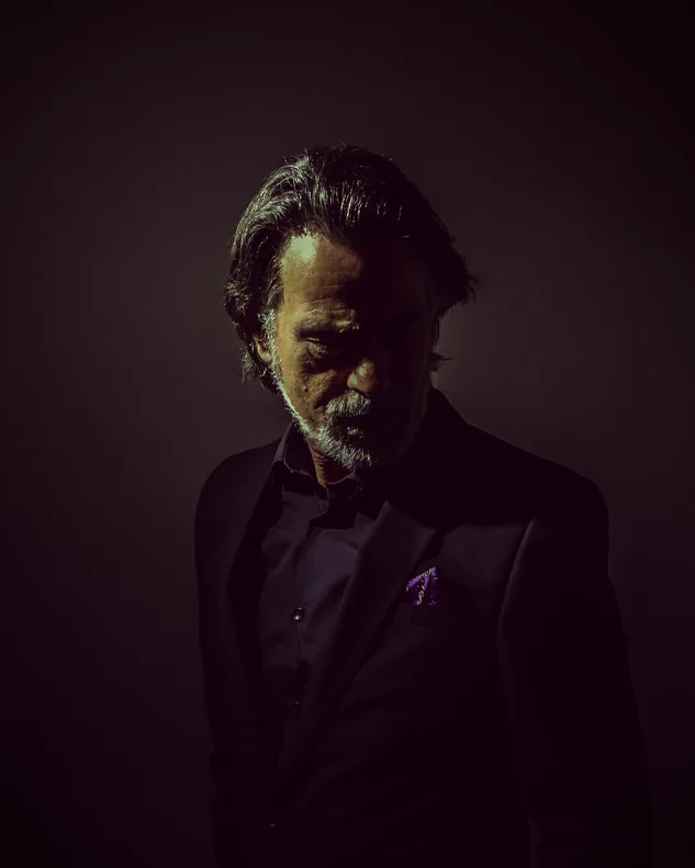A man with grey hair and a beard in a dark suit, low light